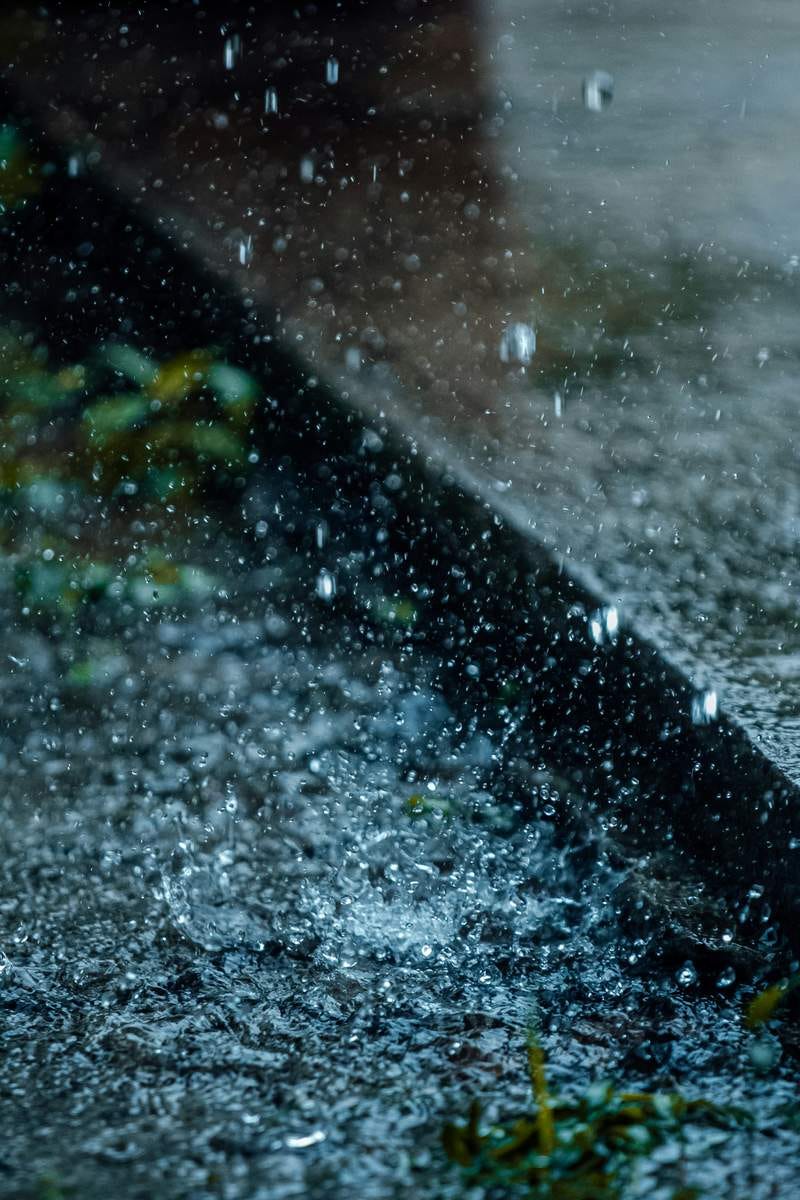 Photo of raindrops pelting down onto a concrete and tarmac roadside.