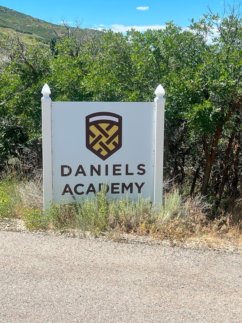 A sign with red text and a red and yellow logo on a white background, saying "DANIELS ACADEMY," in front of some green shrubs with a tall hill or small mountain in the background