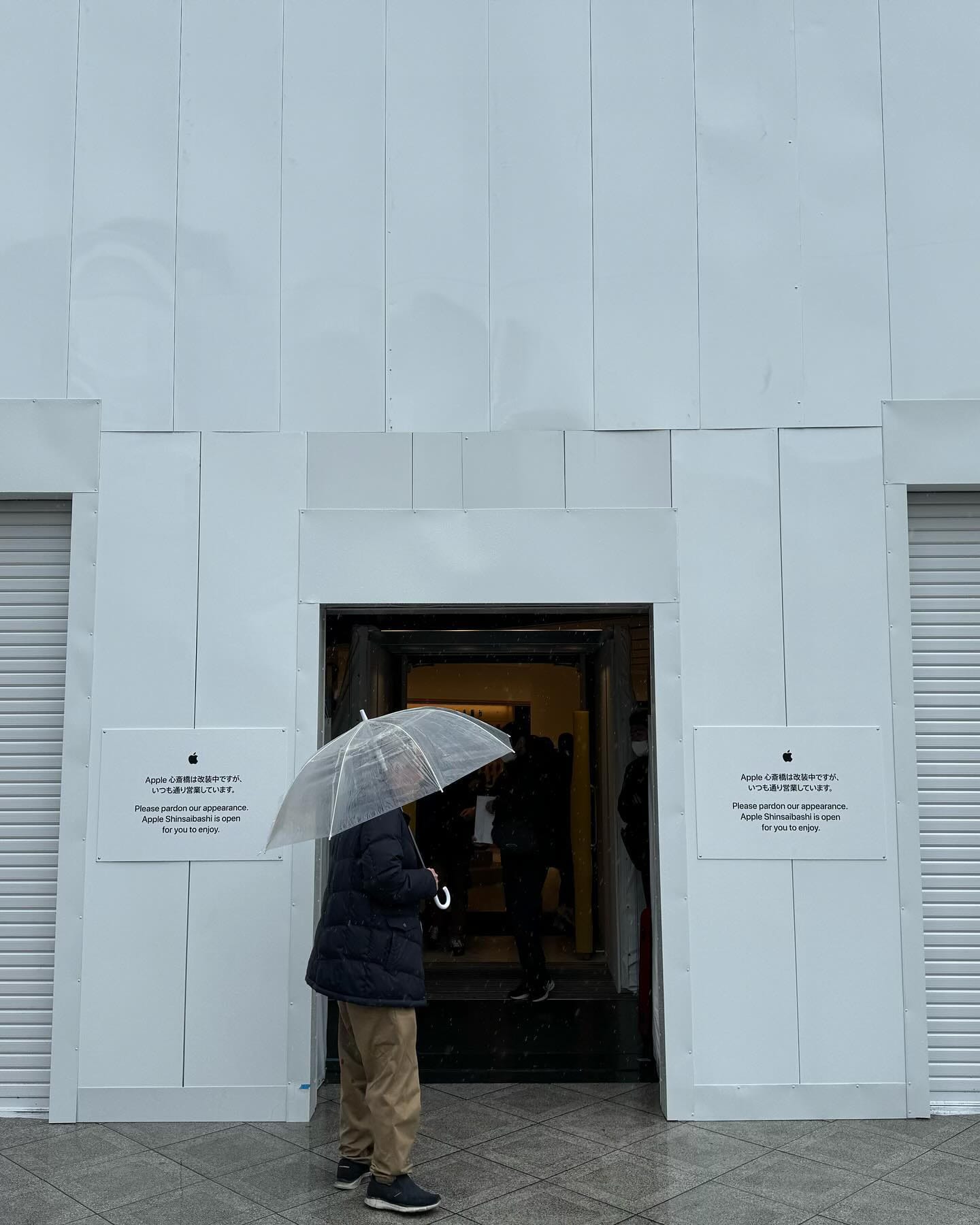 A passerby holding an umbrella passes Apple Shinsaibashi. The facade is covered with construction scaffolding.
