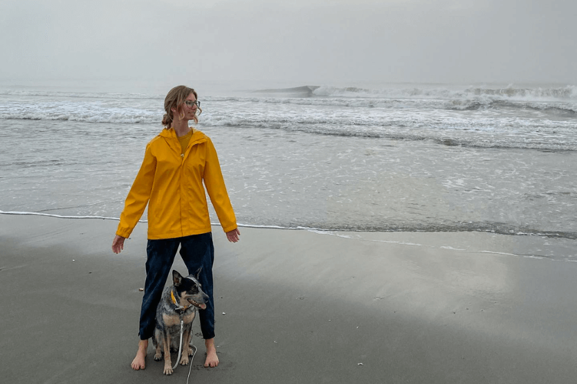 Haley, a blonde woman wearing a bright yellow raincoat, and Scout, her blue heeler in a white collar and leash, stand on the foggy beach at sunrise on the first day of 2023