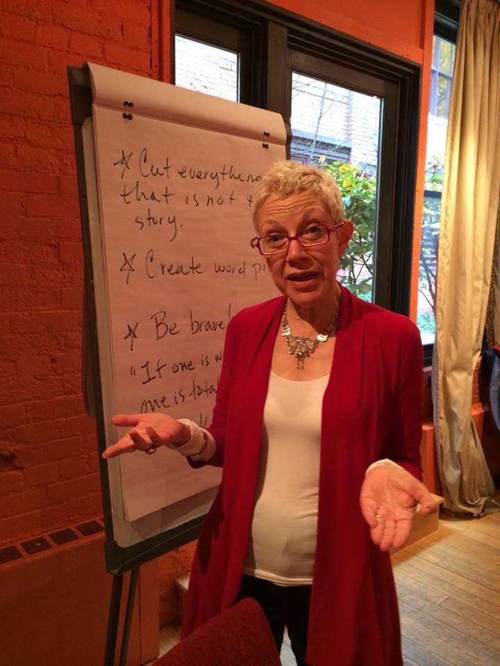 A gray-haired woman in red glasses (Rona Maynard) explains the points on a flip chart.