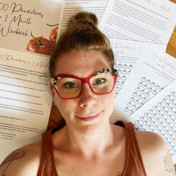 Kate is a white femme with red hair in a bun and she is wearing pink glasses and an orange tank top. She is laying on a wooden floor on top of printed sheets from a workbook.