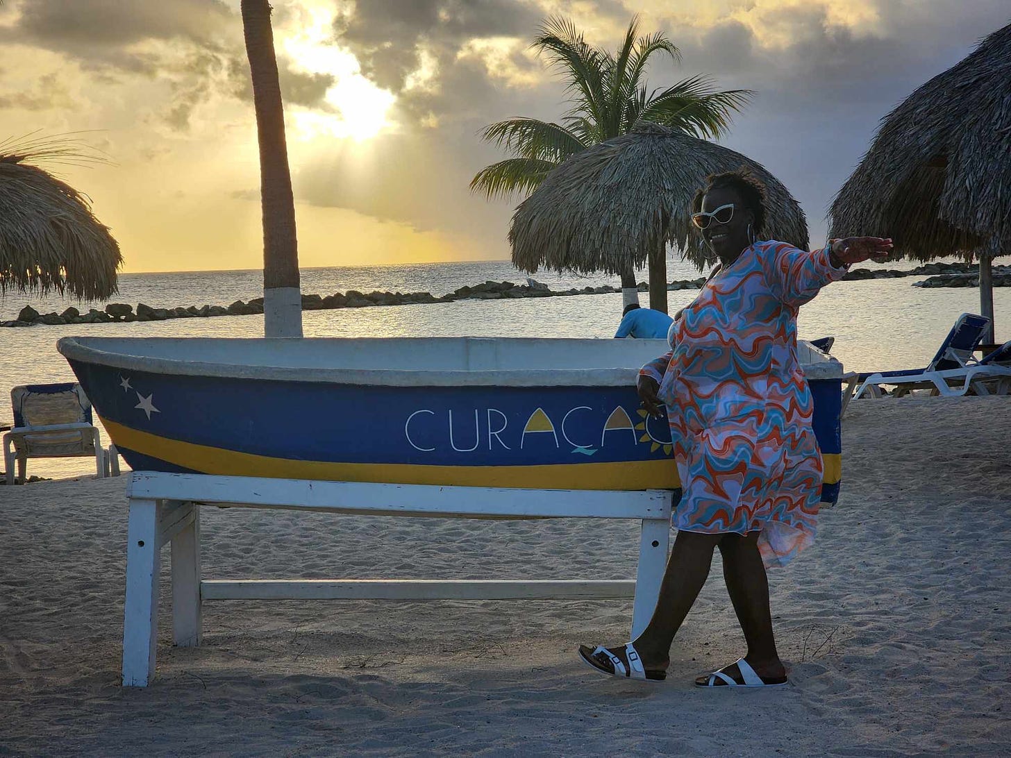 Black woman poses in front a boar that read curacao in blue and yellow. A sunset with palm tree is in the photo's background