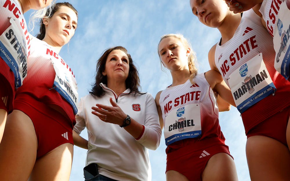 Laurie Henes, wearing a white long-sleeved top with an NC State logo, stands in a huddle with members of NC State's cross country team.