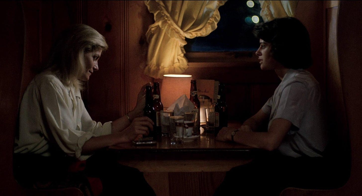 two white women, a blonde and a brunette, sit opposite each other at a dimly lit restaurant booth with bottles of beer on the table in front of them, both look a bit nervous.