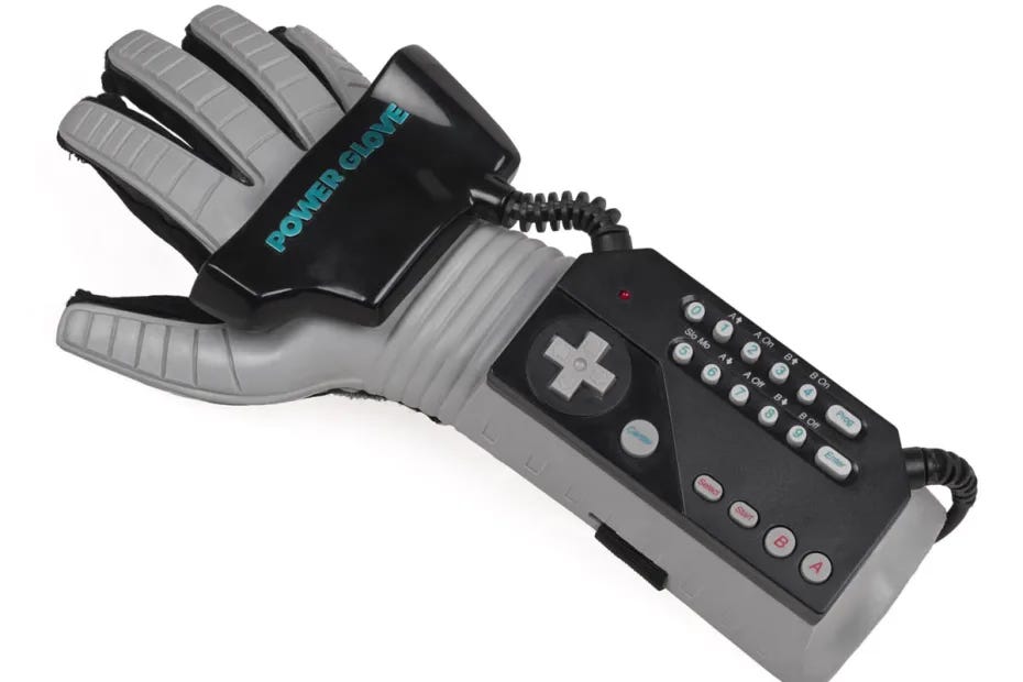 Photograph of the Mattel Power Glove. The Power Glove is a gray plastic gaming controller worn like a glove on the forearm and hand. It features a bulky gray forearm unit with 17 controller buttons and a numeric keypad. The glove itself has a metallic-looking mesh fabric with exposed finger tips and prominent sensors across the knuckles. A thick wire extends from the forearm unit for console connection. This distinctive Nintendo accessory from 1989 has a retro-futuristic, cyberpunk aesthetic.