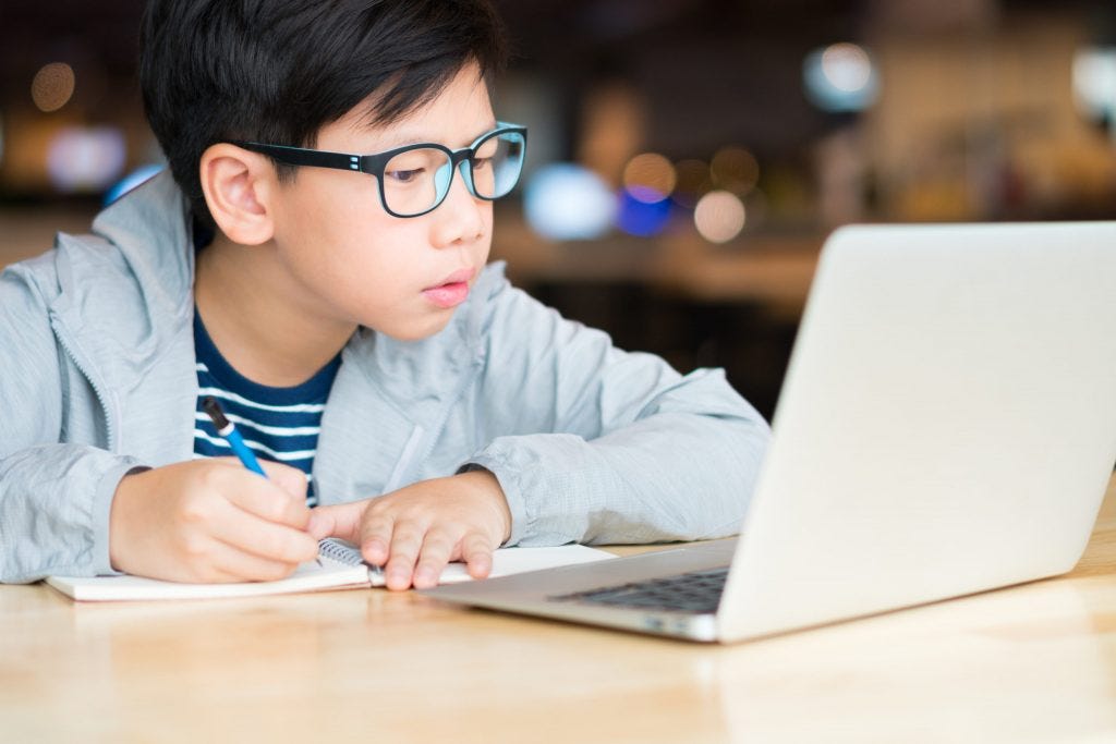 Smart looking Asian preteen boy writing and using computer laptop studying online lessons ...