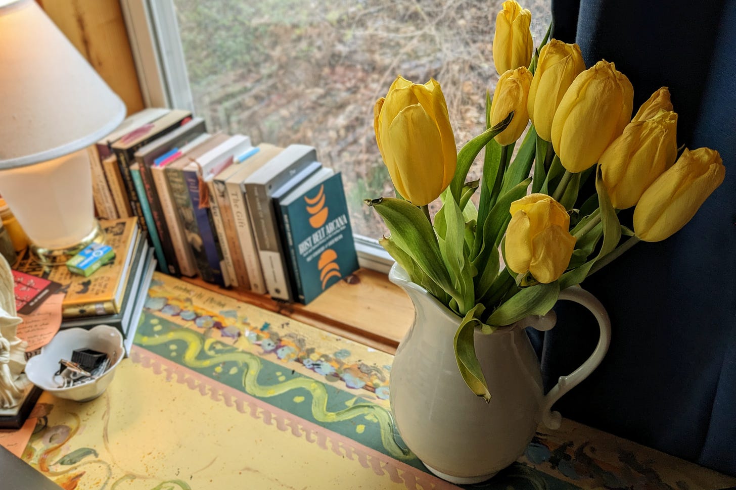 Yellow tulips in a cream-colored vase sit on a colorful desk that's been handpainted. In the background, a row of books rests on the window sill.