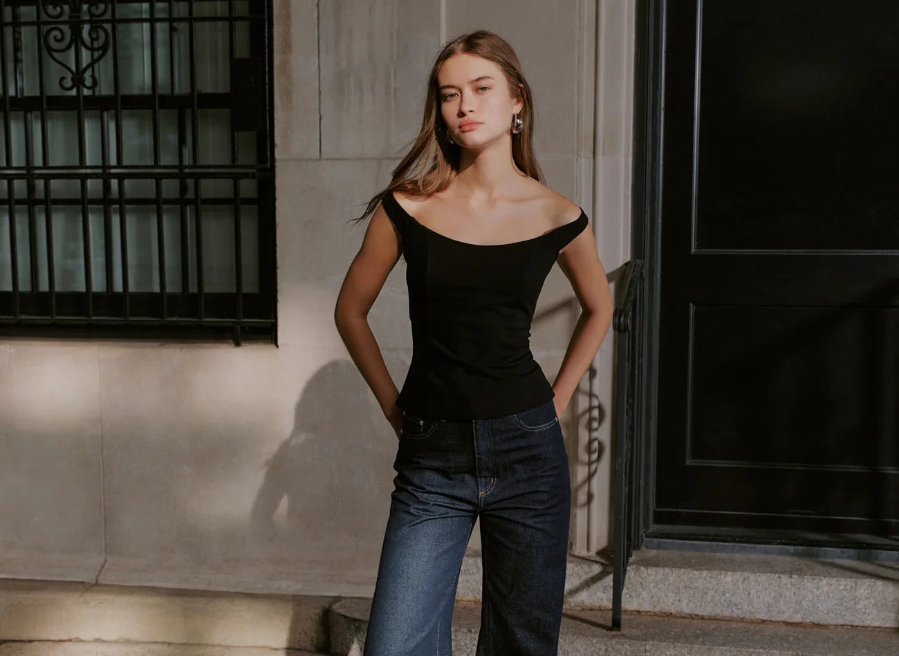a model with dark blond hair stands in front of a concrete building with wrought iron accents. she wears a black off shoulder top with dark wash jeans.