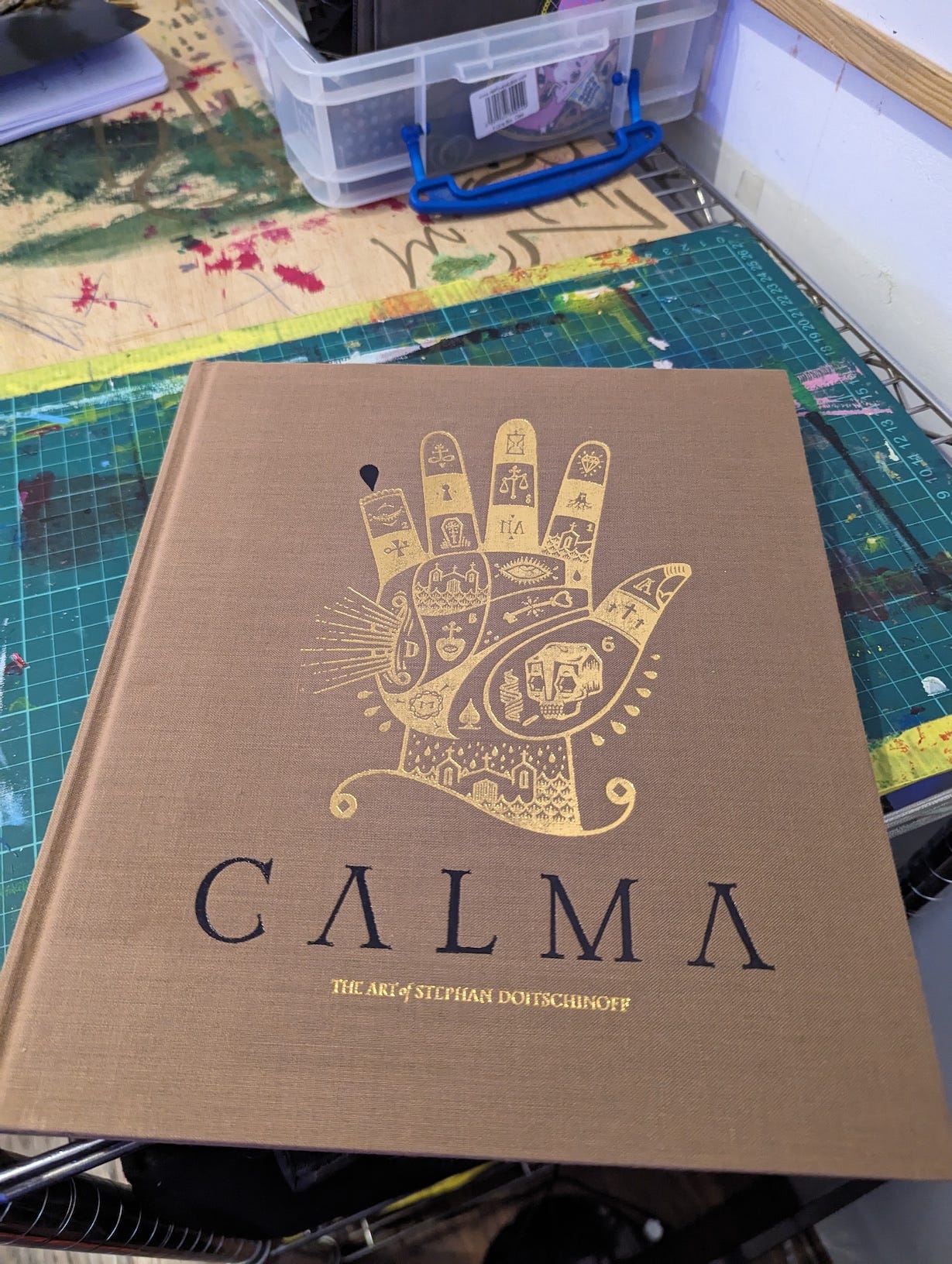 The cover of Calma: The Art of Stephan Doitschinoff features an intricate gold-printed illustration on a brown cloth cover. The artwork depicts an ornate hand filled with symbols, such as an eye, houses, and other mystical imagery. The title "CALMA" and the subtitle "The Art of Stephan Doitschinoff" are prominently displayed in black and gold. The book is set on a workbench, surrounded by art supplies.