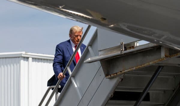 Former President Donald J. Trump, wearing a blue suit with a white shirt and red tie, is pictured with blue sky and a white building behind him as he boards a plane.