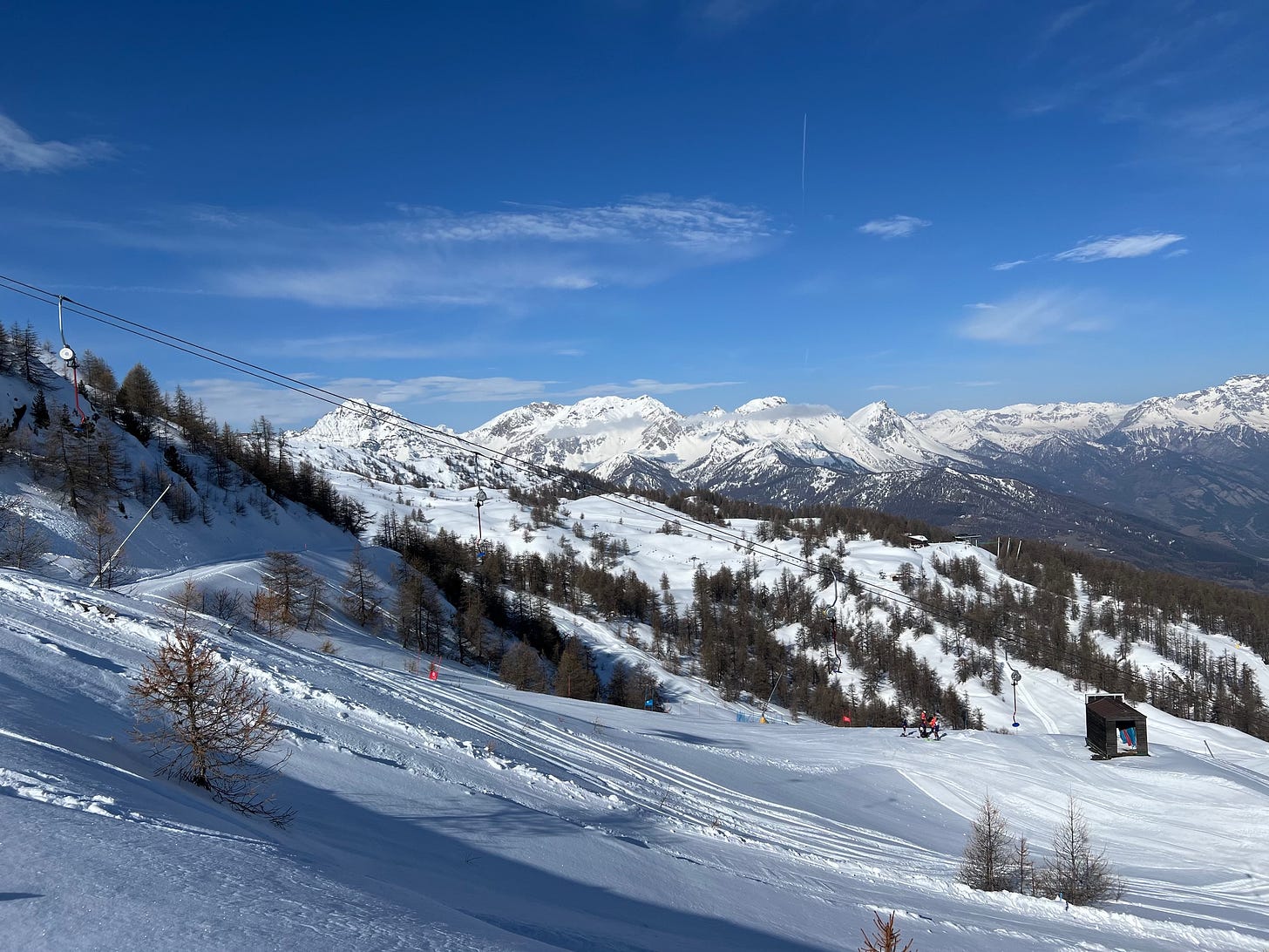 A scenic view of a snowy mountain landscape with ski slopes and scattered pine trees. A ski lift runs diagonally across the scene, with skiers visible near a small wooden hut on the right. In the background, rugged, snow-covered peaks rise under a bright blue sky with scattered clouds.