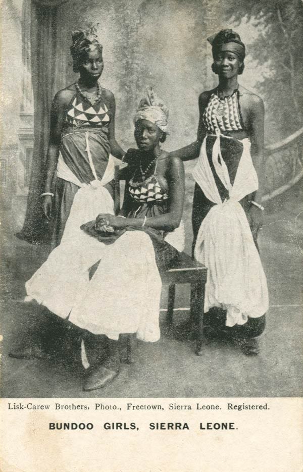 An image of three young black women, two are standing on either side of the seated woman wearing long skirt posing for the picture. The woman standing on the right is smiling, whereas the other two women have a straight face.