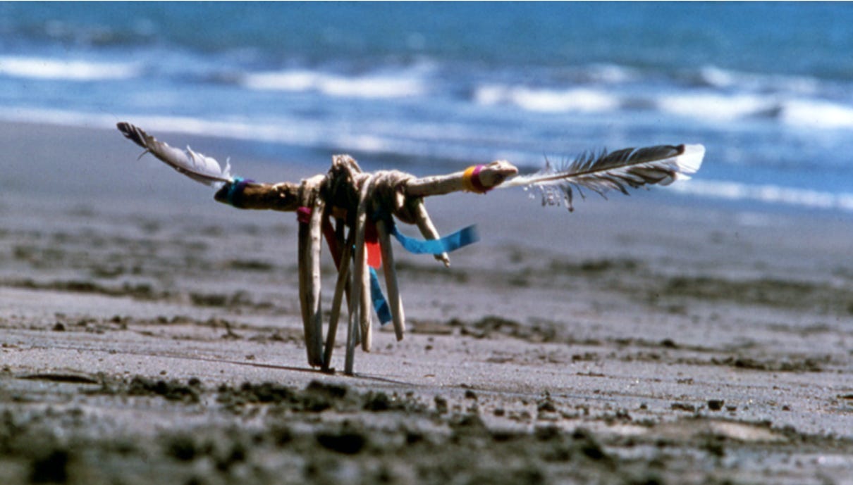 An object set into the sand with the ocean in the distance. The object is small, made of twigs, bits of fabric, and two feathers like arms outstretched.