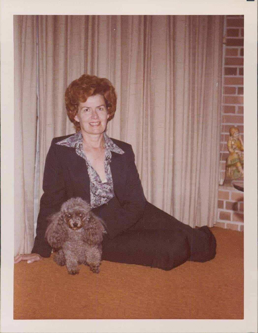 A women in a blue suit with red hair sitting on the floor with her toy poodle