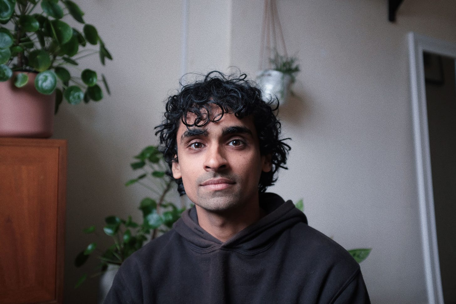 This is a headshot of Neef, who is seated in front of some indoor green plants. He has brown skin, dark-brown eyes, and short black curly hair. He's wearing a brown-grey hoodie sweater. He's staring directly into the camera with his mouth closed, and looks relaxed.
