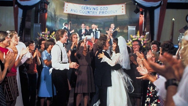 A newly married couple surrounded by clapping friends, a band, and a banner that says "Serving God And Country Proudly" from Michael Cimino's The Deer Hunter.