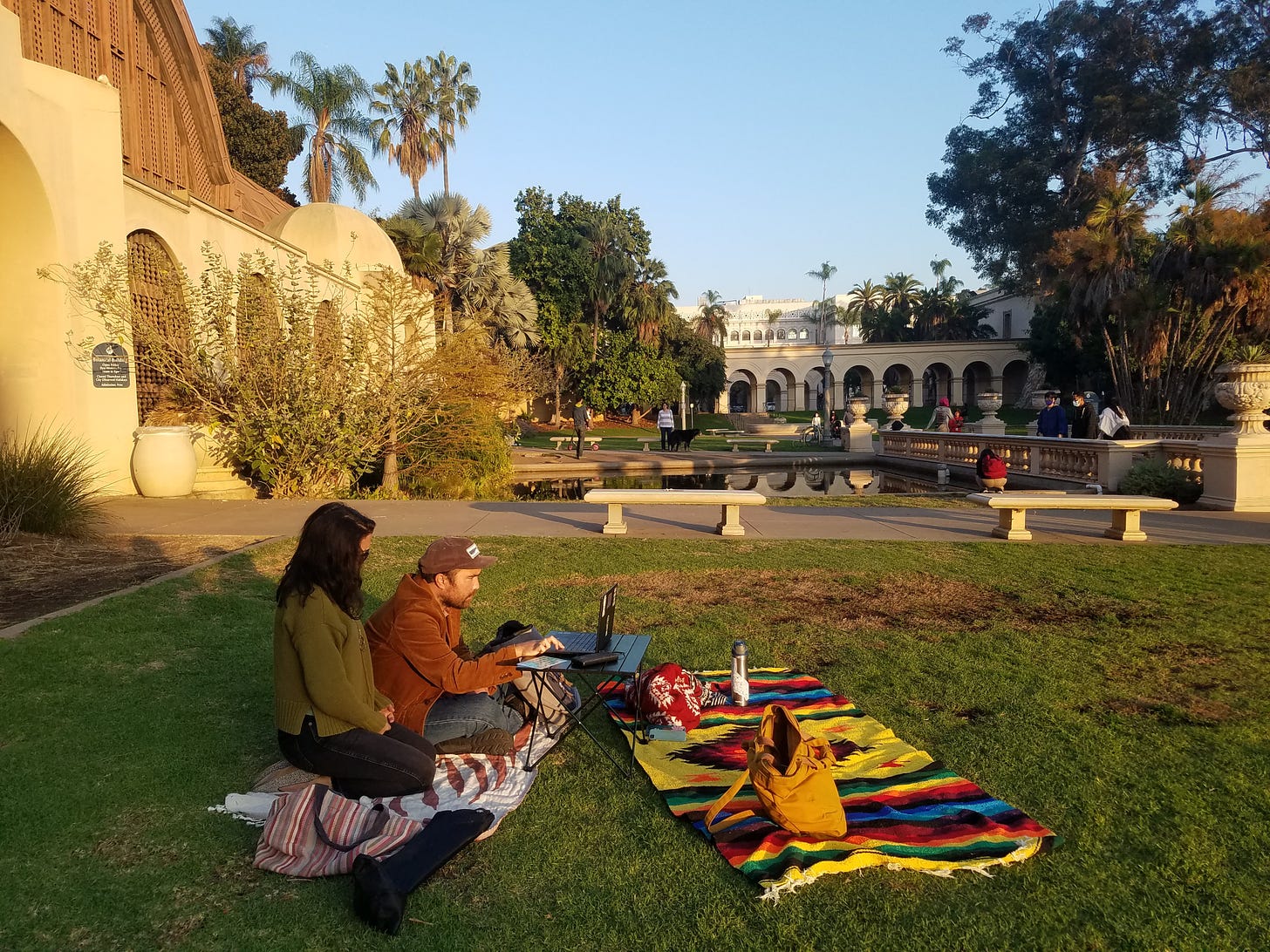 A woman and man, sister and brother, sit together on a blanket on a lawn. Behind them is a stately building and a pond, all illuminated in late afternoon golden light. The man is looking at a computer on a small table in front of them.