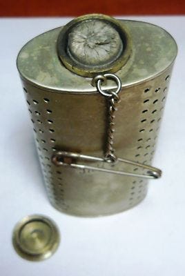 A photograph of a metal canister somewhat like a hip flask but taller. It has rows of holes down each side and a safety pin attachment for fastening it to the wearer's clothing.