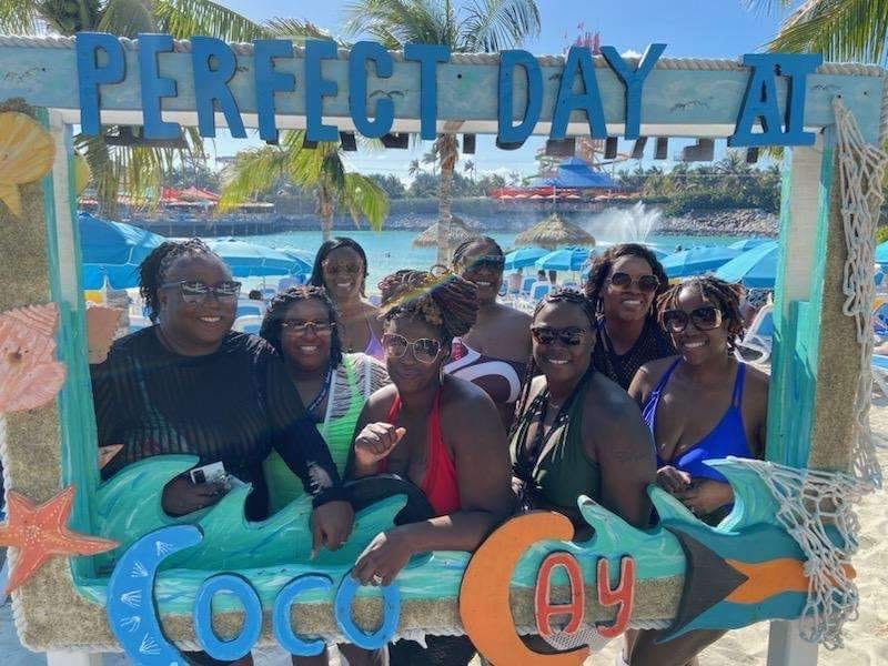 8 Black women posing in front of wooden frame that reads "Perfect Day at Coco Bay"