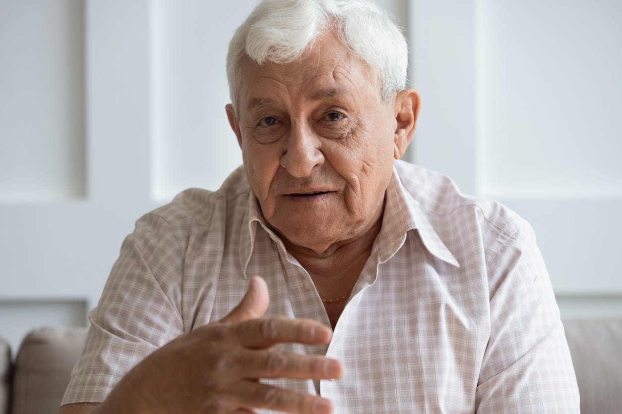 As if seriously speaking toward the camera, older, white man with full head of gray hair, morions with his right hand.