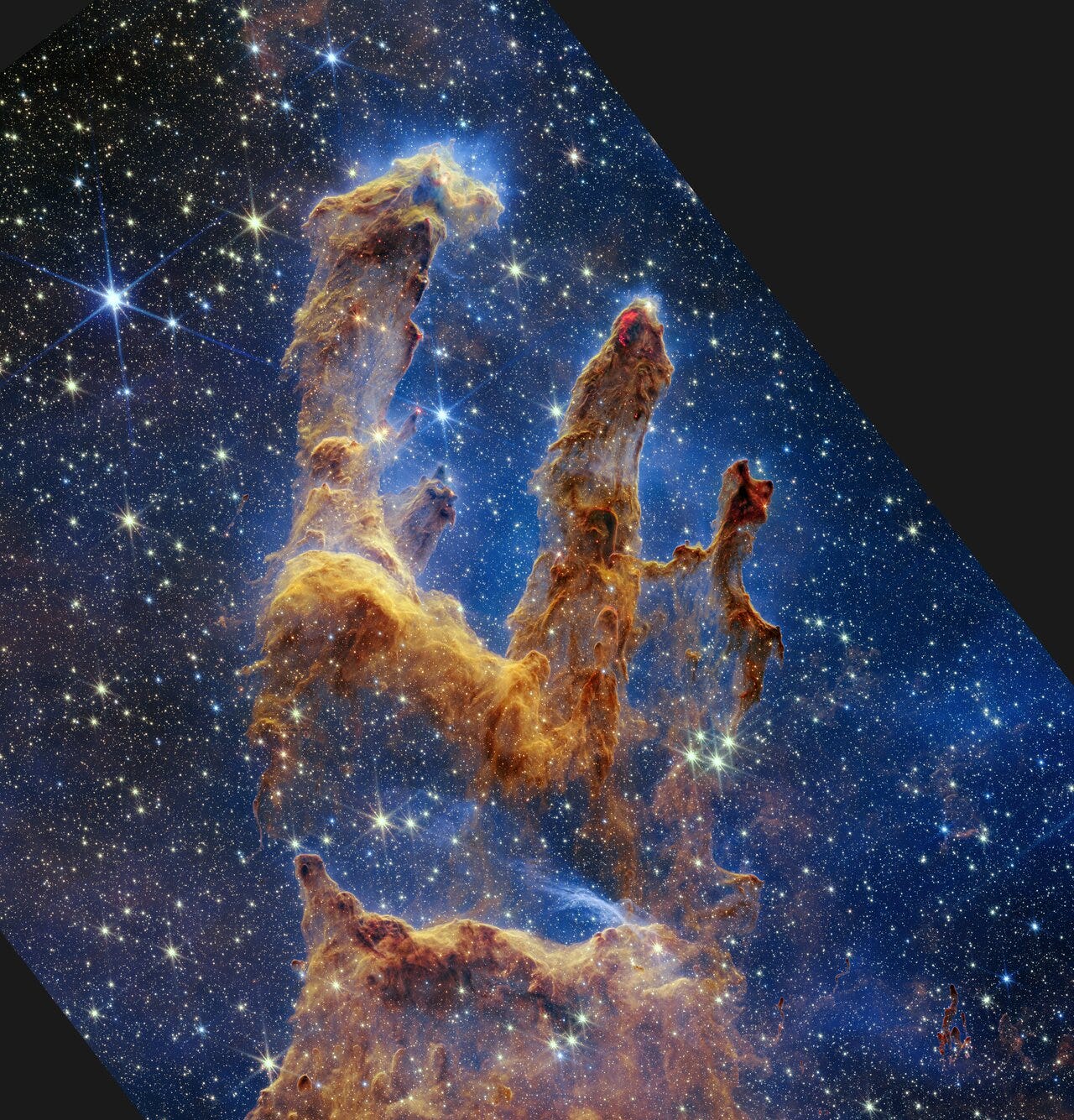 Pillars of reddish-brown interstellar gas appear as "fingers" reaching up against a dark blue background dotted with yellow, blue, and yellow-green stars. 