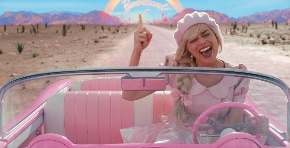 A blonde woman, wearing pale pink clothes, sings as she drives a bubble-gum pink convertible car long a road in a desert landscape with a backdrop of mountains and a blue sky with a rainbow under which script lettering reads 'Barbieland'.