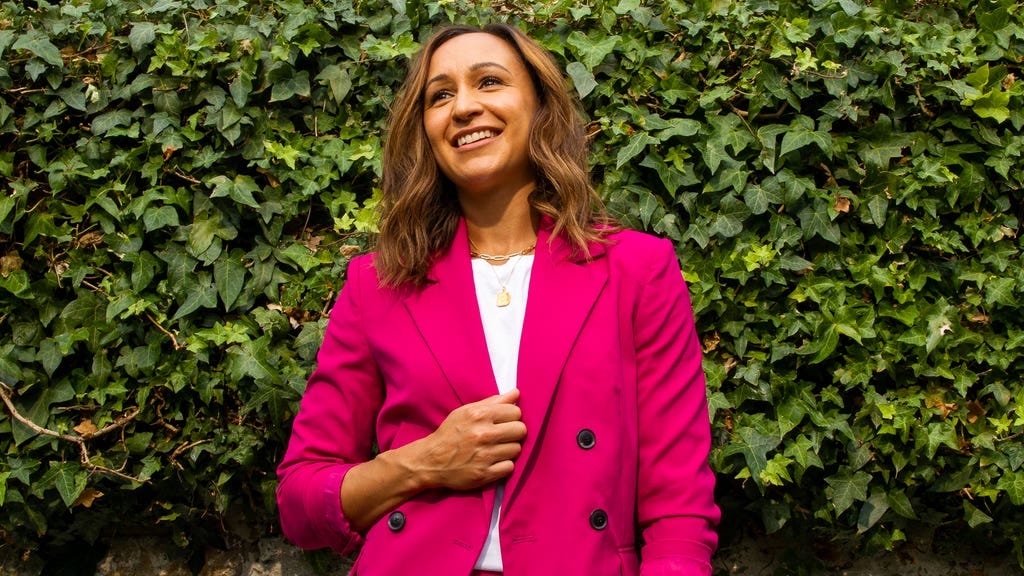 A portrait shot of Jessica Ennis-Hill wearing a hot pink trouser suit, standing in front of a wall covered in green ivy