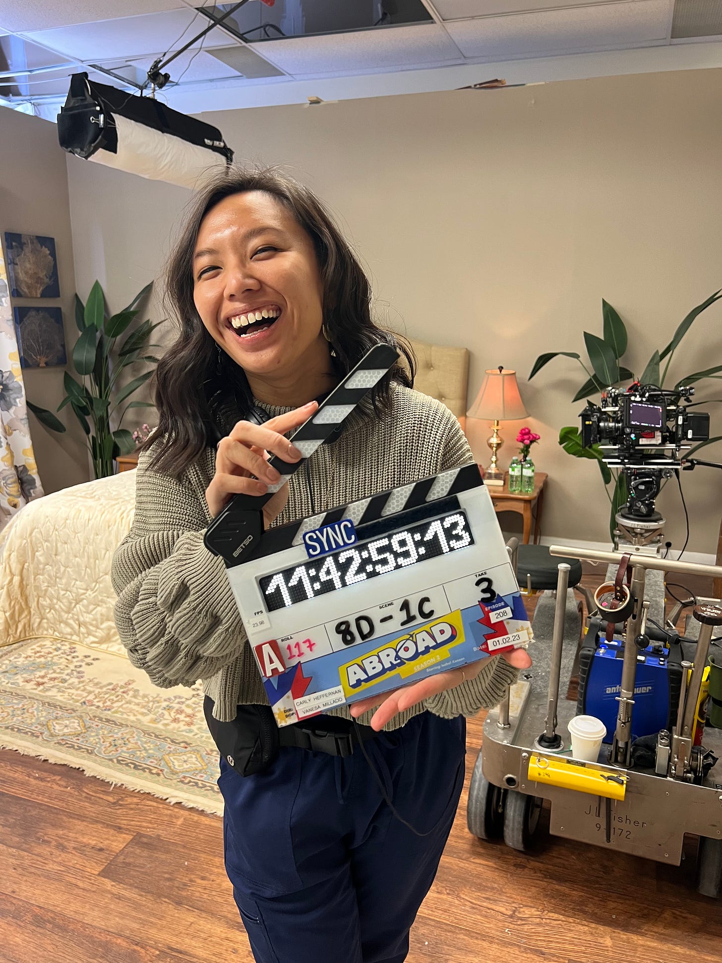 Justine standing on set of a TV show holding a clapper that says ABROAD. There is a couch and camera behind her.