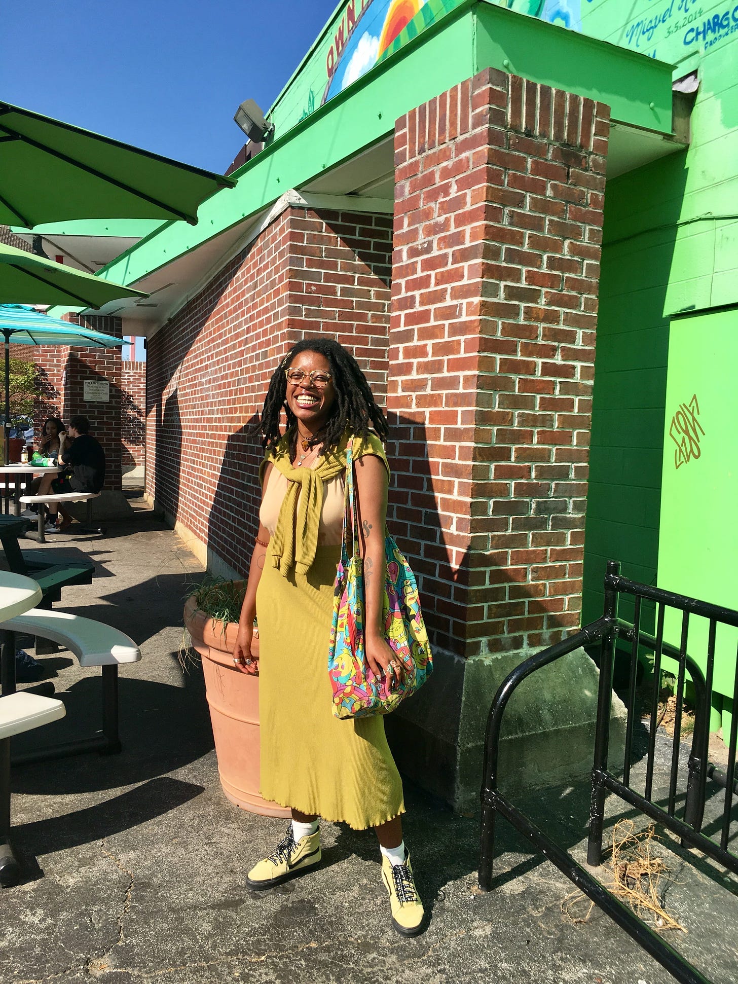 a young Black woman smiles while standing in front of a brick building. it’s a healthy food store with a green awning. the woman wears a tan bodysuit with a green skirt set. a matching green sweater is tied around her shoulders and she carries a large colorful tote bag. she wears yellow ATCQ Vans shoes.