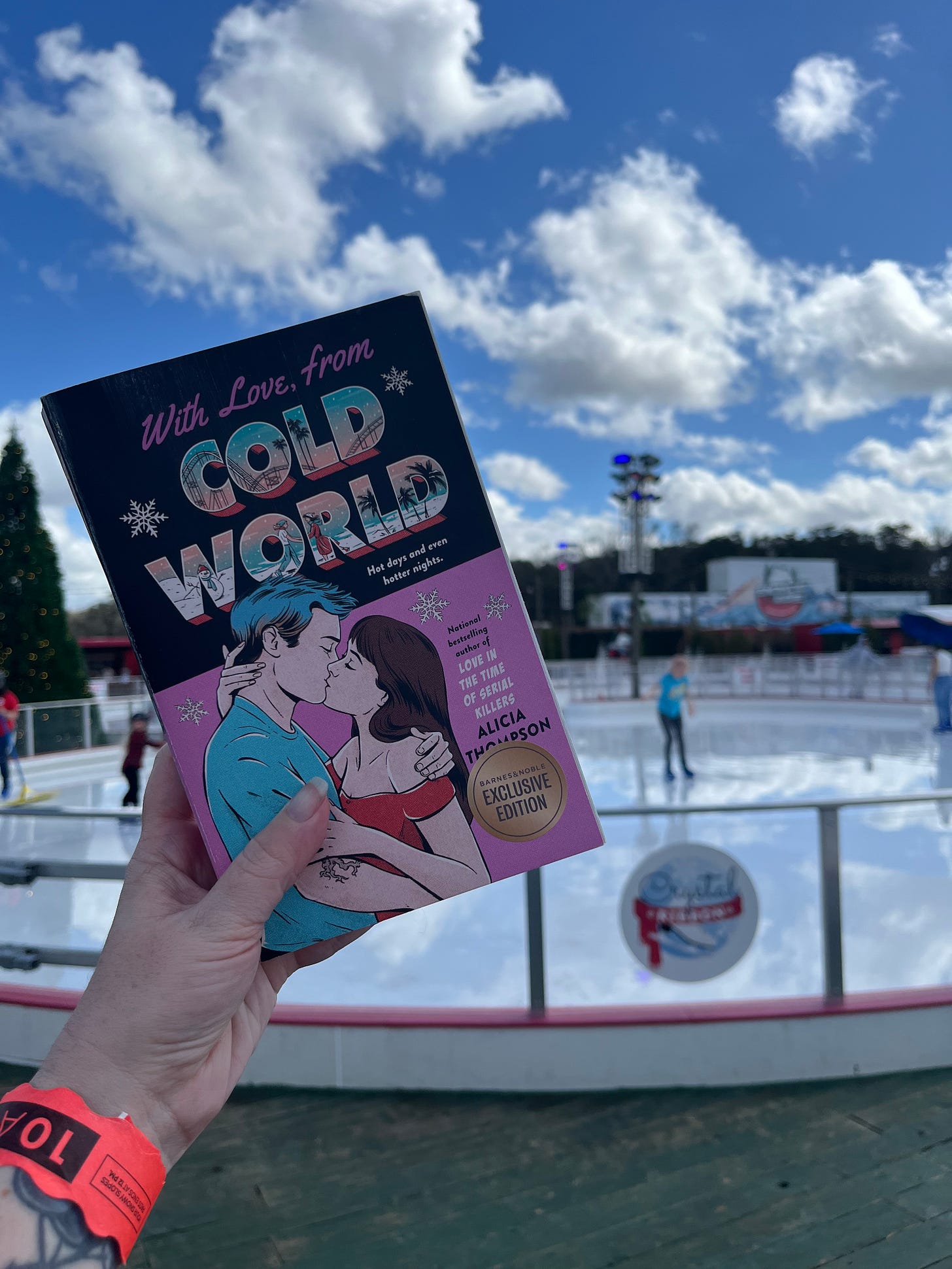 Picture of my hand holding up the pink cover copy of WITH LOVE, FROM COLD WORLD in front of the ice skating rink