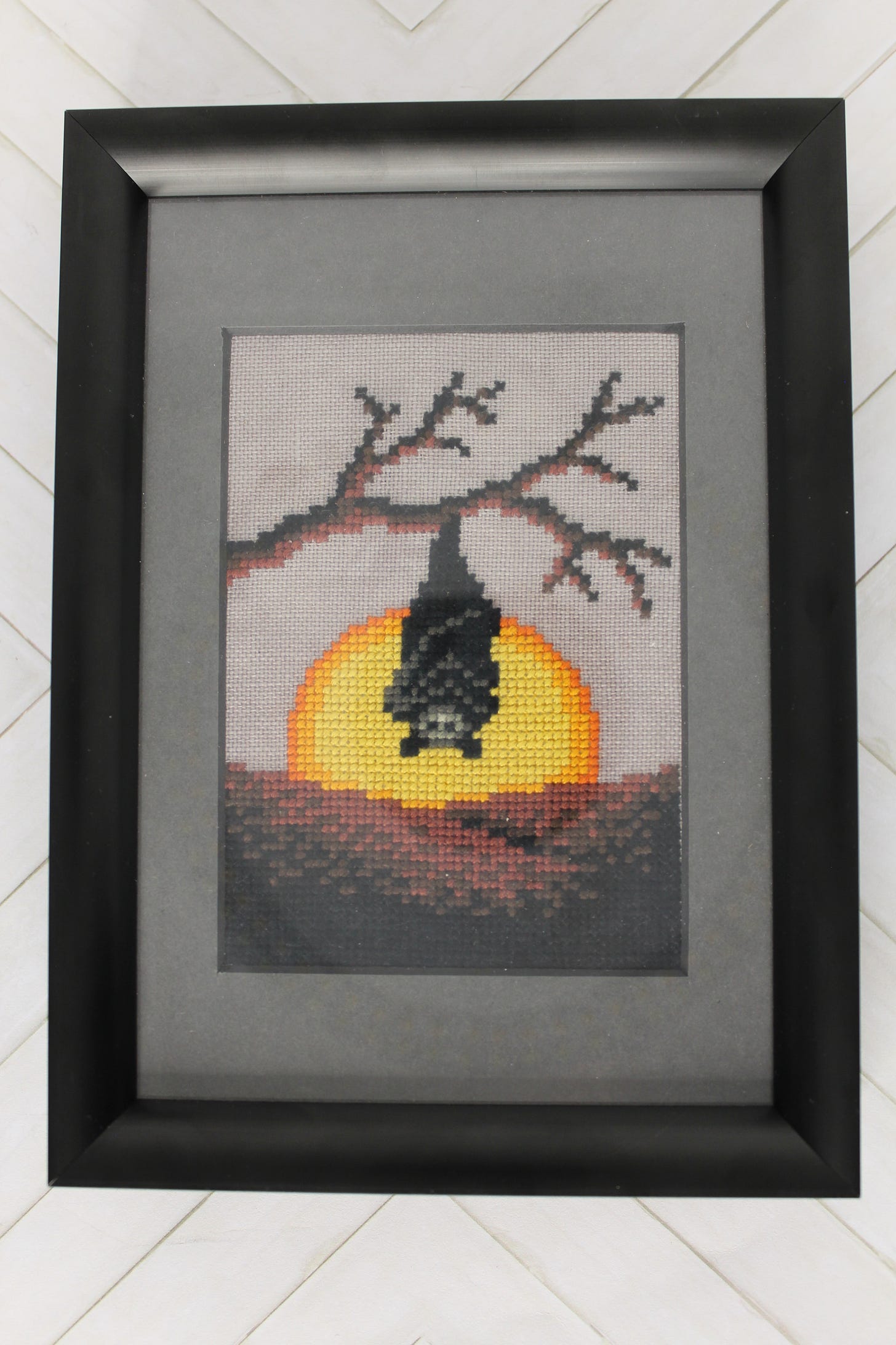 A framed and matted cross stitch pattern of a bat hanging by its feet from a tree branch.