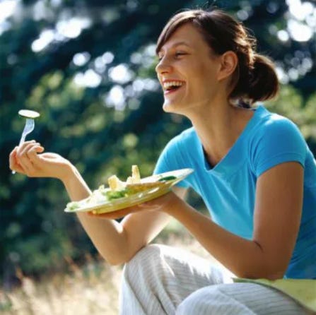 White woman in a blue tshirt sitting outside with a bowl of salad and laughing