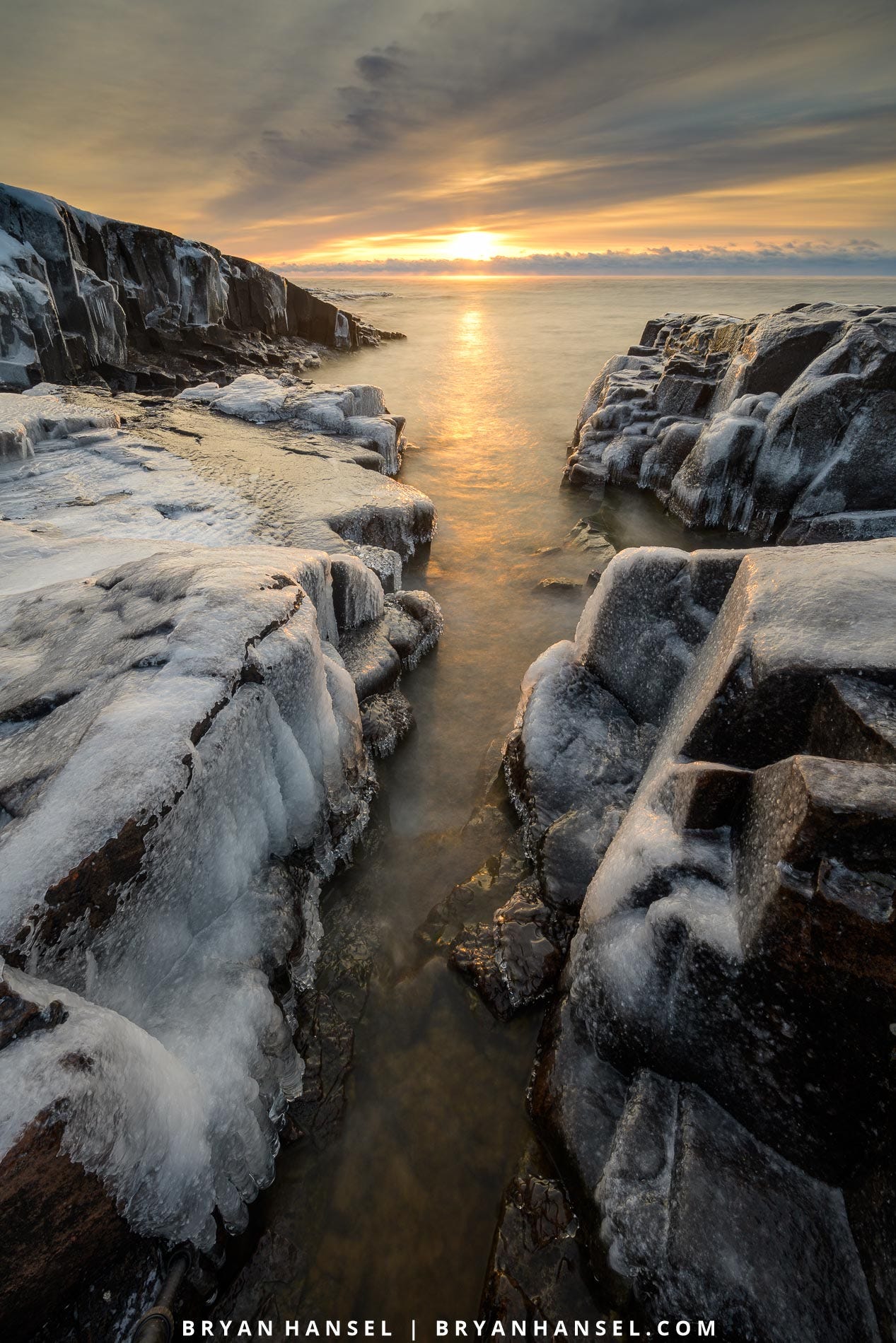 A photo of sunrise coming directly down a slot canyon in a basalt shoreline on Lake Superior. Sunrise is yellow and it is making the ice on the basalt glow yellow.