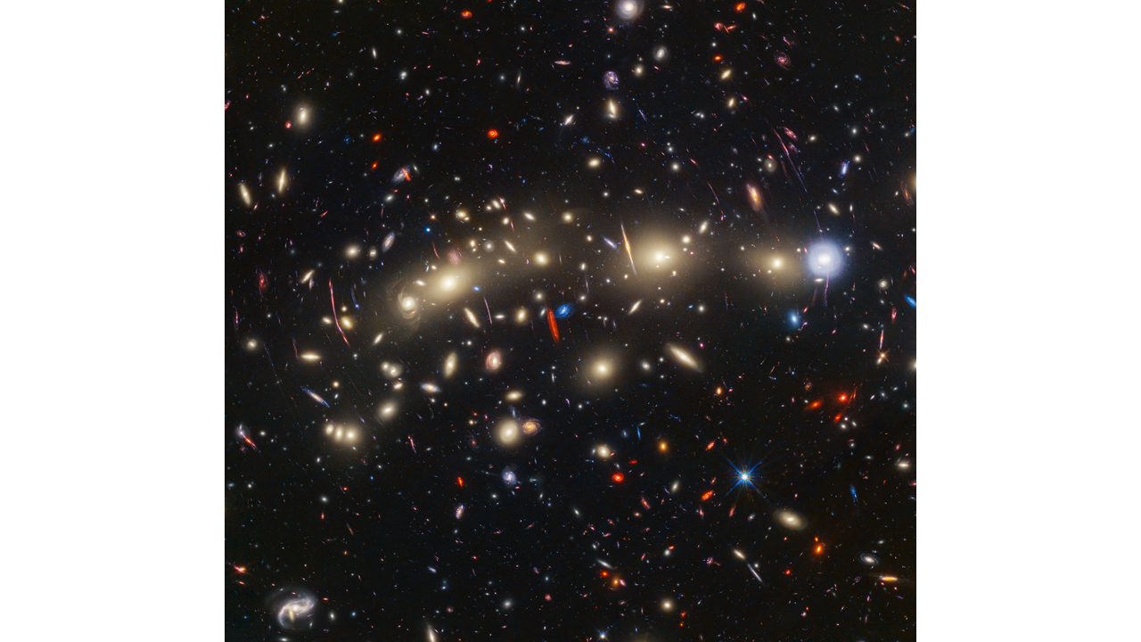 A field of galaxies on the black background of space. In the middle, stretching from left to right, is a collection of dozens of yellowish spiral and elliptical galaxies that form a foreground galaxy cluster. They form a rough, flat line along the center. Among them are distorted linear features, which mostly appear to follow invisible concentric circles curving around the center of the image. The linear features are created when the light of a background galaxy is bent and magnified through gravitational lensing. At center left, a particularly prominent example stretches vertically about three times the length of a nearby galaxy. A variety of brightly colored, red and blue galaxies of various shapes are scattered across the image, making it feel densely populated. Near the center are two tiny galaxies compared to the galaxy cluster: a very red edge-on spiral and a very blue face-on spiral, which provide a striking color contrast.