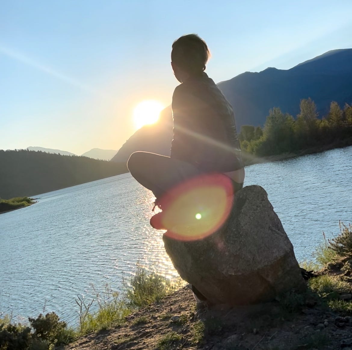 Lyric sits on a log, overlooking a lake and beautiful mountains as the sunsets.