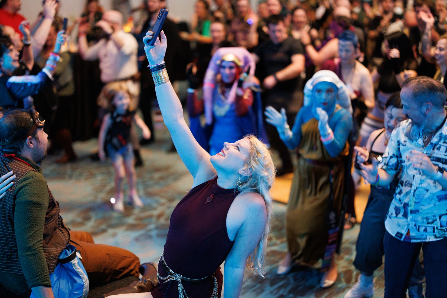 Cass in a maroon, halter-necked formal gown and lots of gold jewelry, grinning widely with cell phone held high in the air as she bends backwards to try to get a room full of partygoers in frame