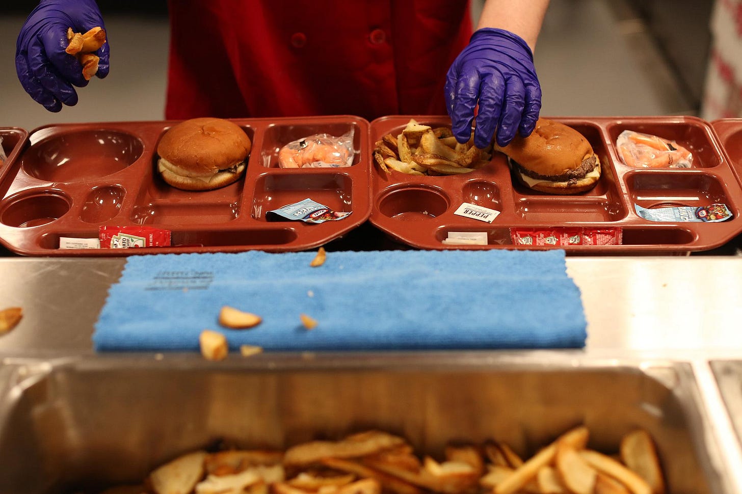 An incarcerated person wearing food preparation gloves adds French fries to a food tray that contains a hamburger and a small bag of baby carrots.  