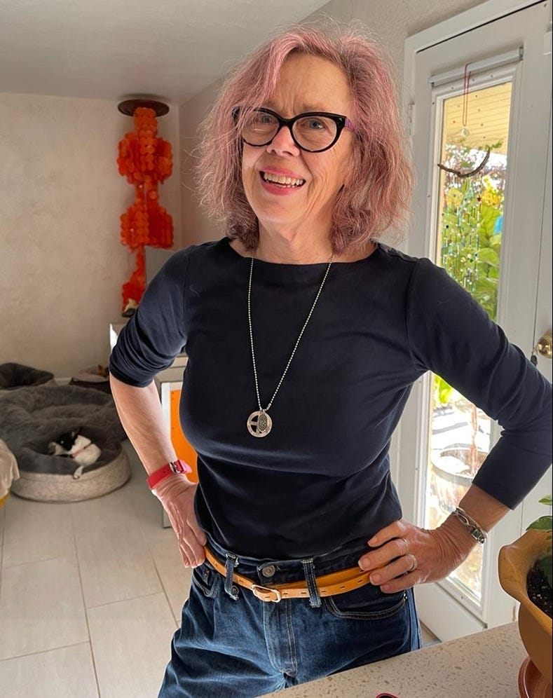 photo of Margaret McInerny, a white woman in her 70s with shoulder-length pink hair and eyeglasses. she's wearing a black shirt and jeans and is posing with her hands on her hips