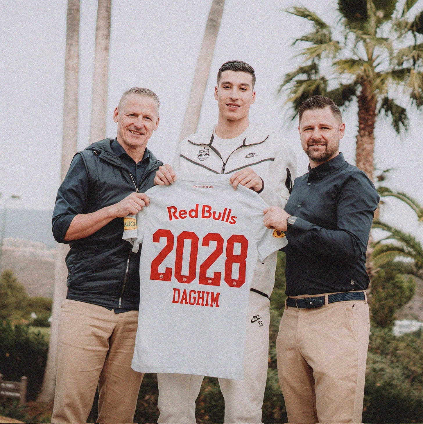 A photo of Adam Daghim holding a white Red Bull Salzburg shirt with '2028 Daghim' on the back. He's stood between two Salzburg executives wearing green tops and beige trousers. In the background are palm trees.