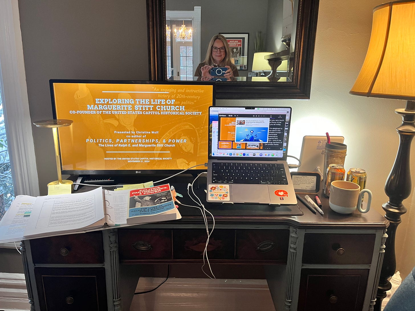 A blonde woman takes a selfie in front of a mirror which hangs above a desk with a TV monitor, a laptop, papers, a book, and four beverages