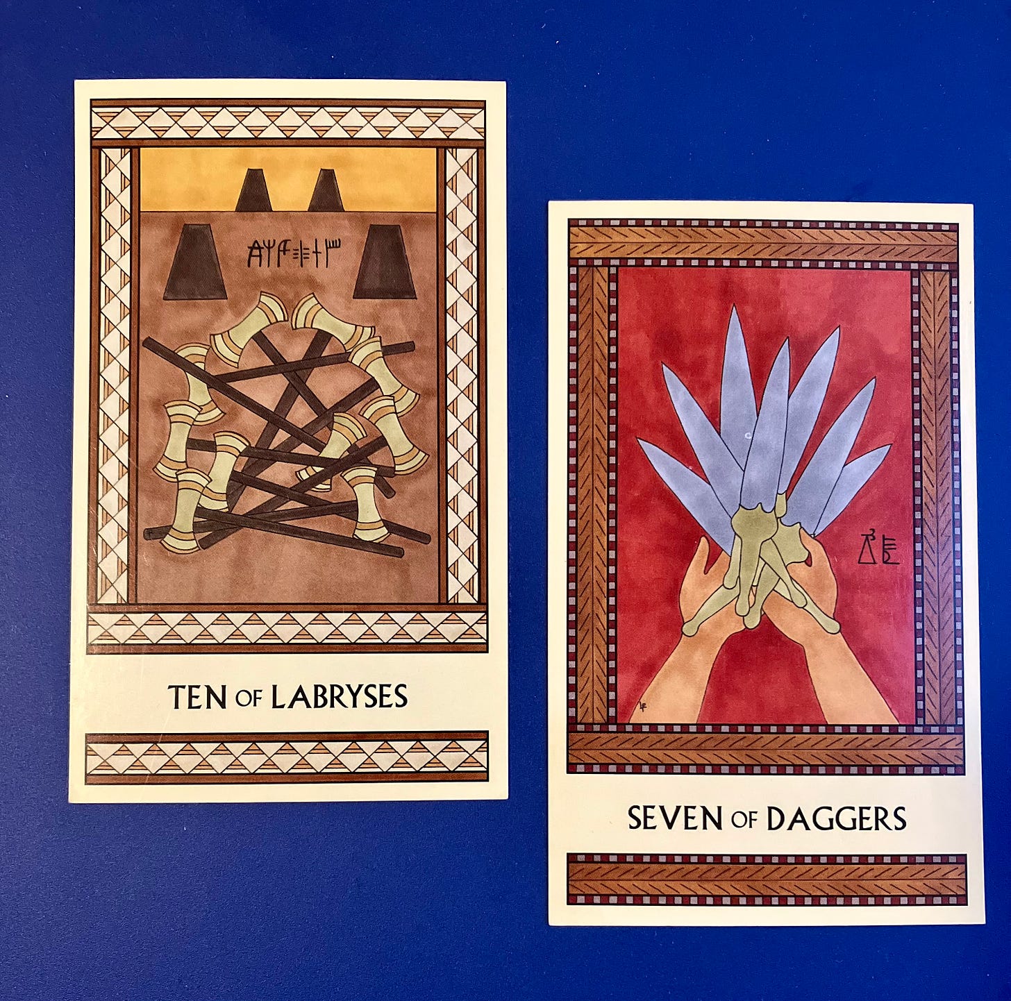 Two Minoan Tarot cards side by side on a deep blue background. The Ten of Labryses is in shades of gold and tan. It shows ten labryses lying in a heap on the floor, with four empty labrys stands in the background. The Seven of Daggers is in shades of deep red and tan. It shows two hands holding a pile of seven daggers all at once.