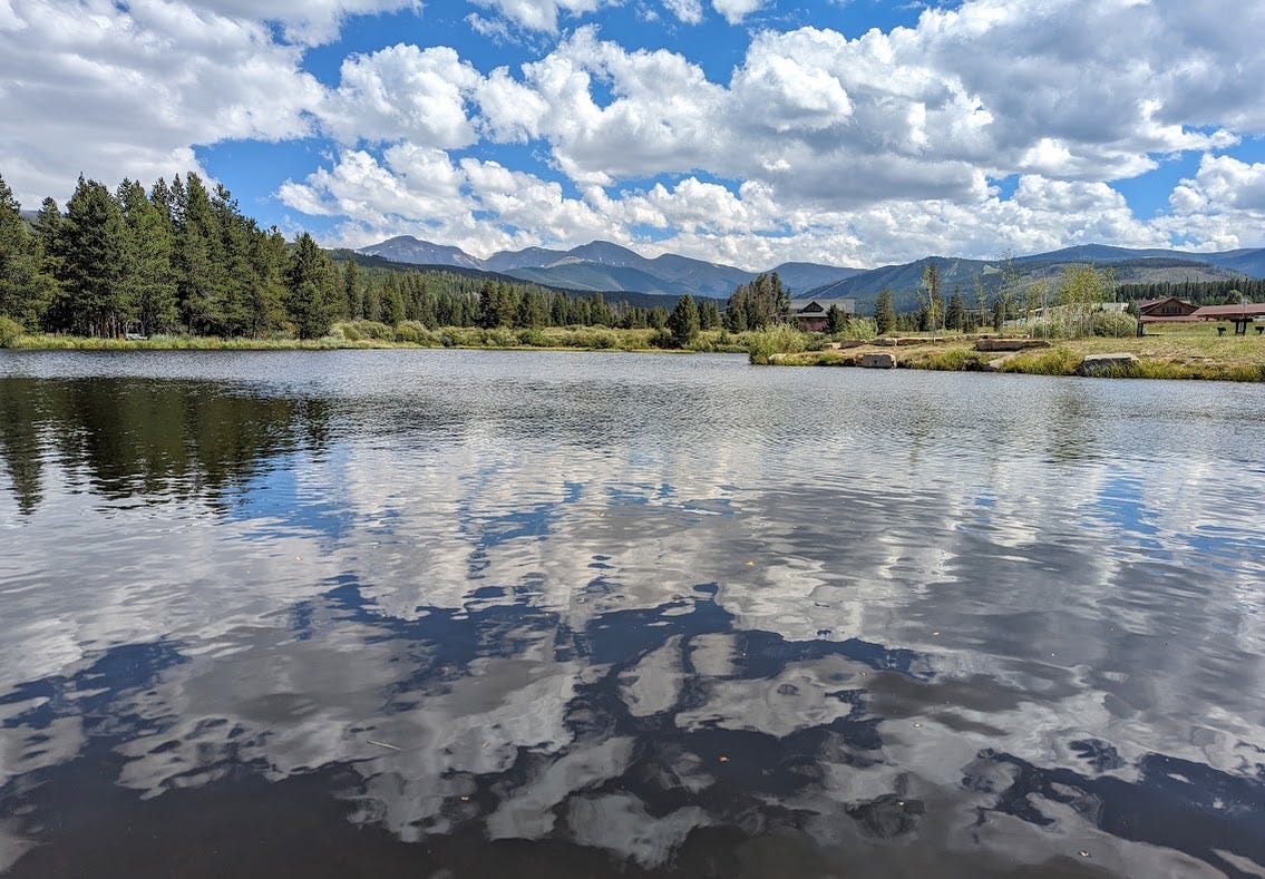 A pond reflecting white clouds, with trees and mountains in the distance
