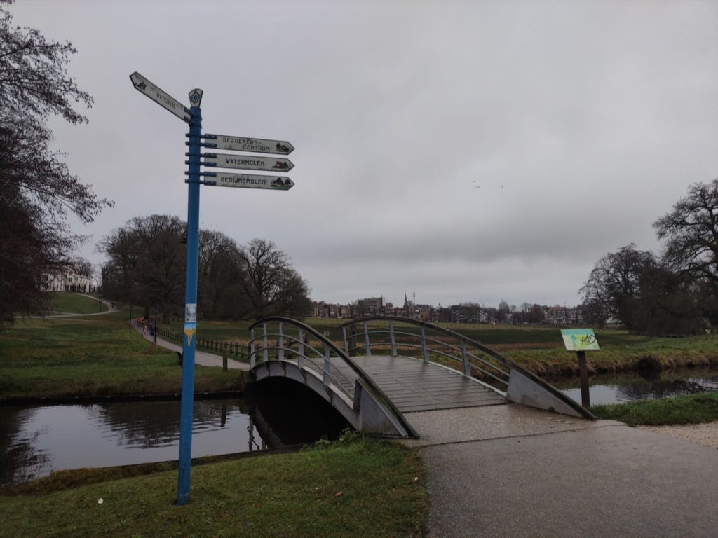 A small humped-bridge over a canal at the end of the downhill section. The paths winds uphill away from the camera, with the white walls of the Stadsvilla visible through the trees.