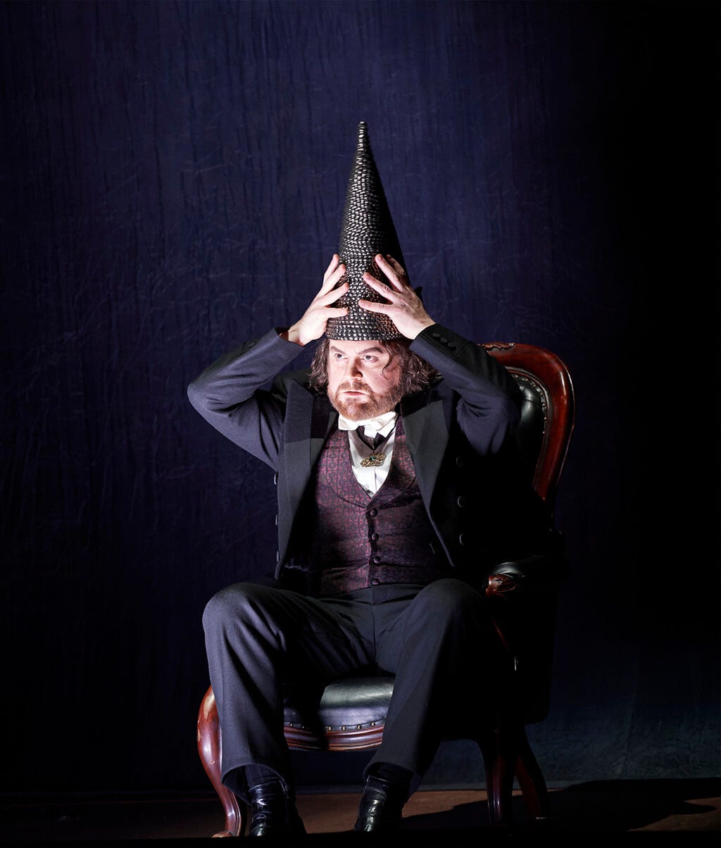 An image from a production of Rigoletto. A man in a Victorian three-piece suit, seated in a chair, places a dunce cap on his head.