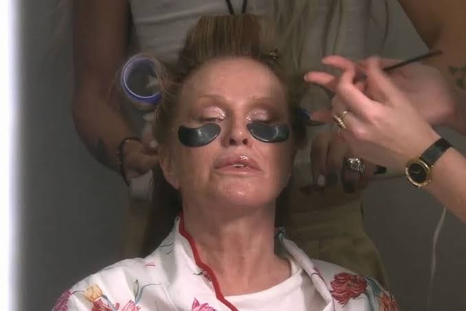 Foto de uma mulher mais velha, loira, com bobs no cabelo, patch pra diminuir olheiras debaixo dos olhos e pessoas em volta dela ajeitando a aparência