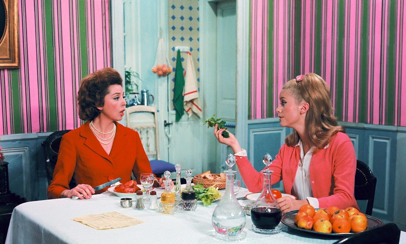 Two women, one older and one younger, both wear red as they sit at a dining table with a lunch spread with fruit, vegetables, bread, and bottles. They are looking at each other while the older woman is speaking and the younger woman holds a leaf of lettuce. The room has blue-painted wood paneling on the lower half and bubble-gum pink and green striped wallpaper on the upper half. The room looks into a kitchen.