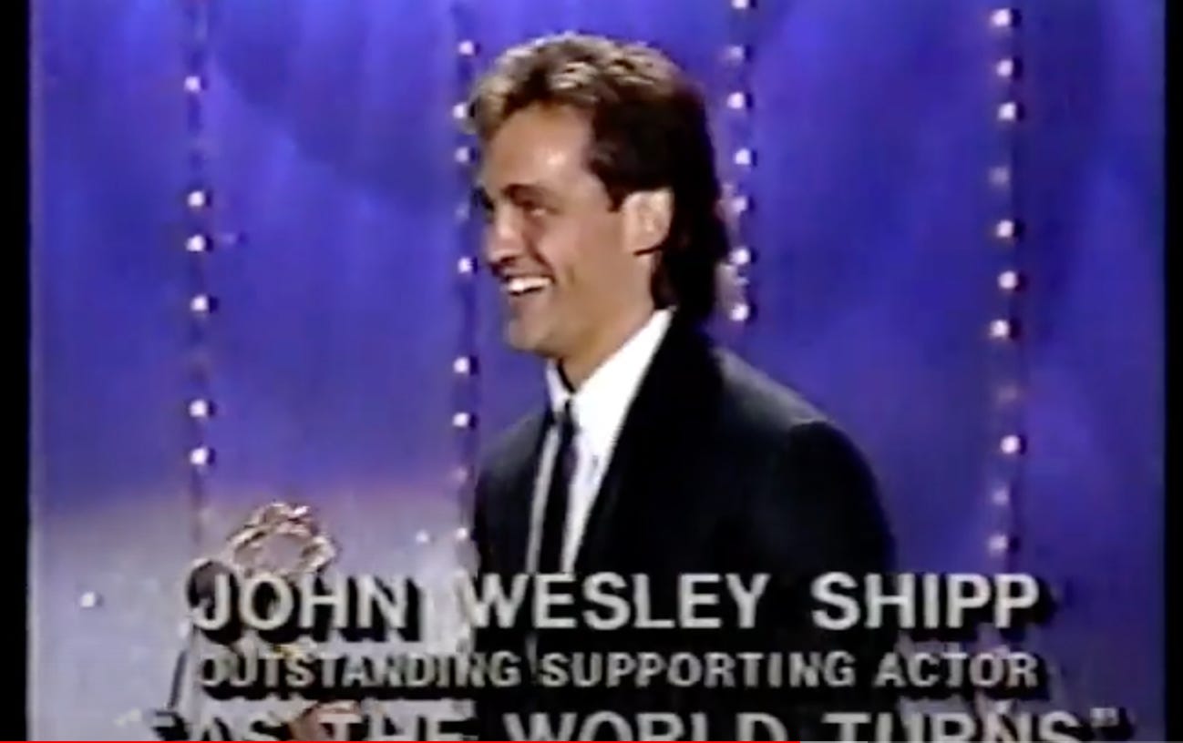 John Wesley Shipp has brown hair and is wearing a black blazer and matching tie. He is holding a Daytime Emmy which has a woman holding the world and is made of gold.