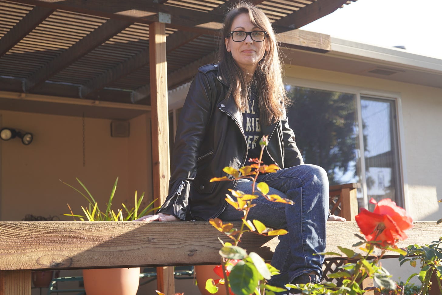 a photo of Kendra, a white woman with brunette hair, sitting on a bench during golden hour. She’s wearing a black jacket and a Bitter Southerner t-shirt that says, “hell or High Water.”