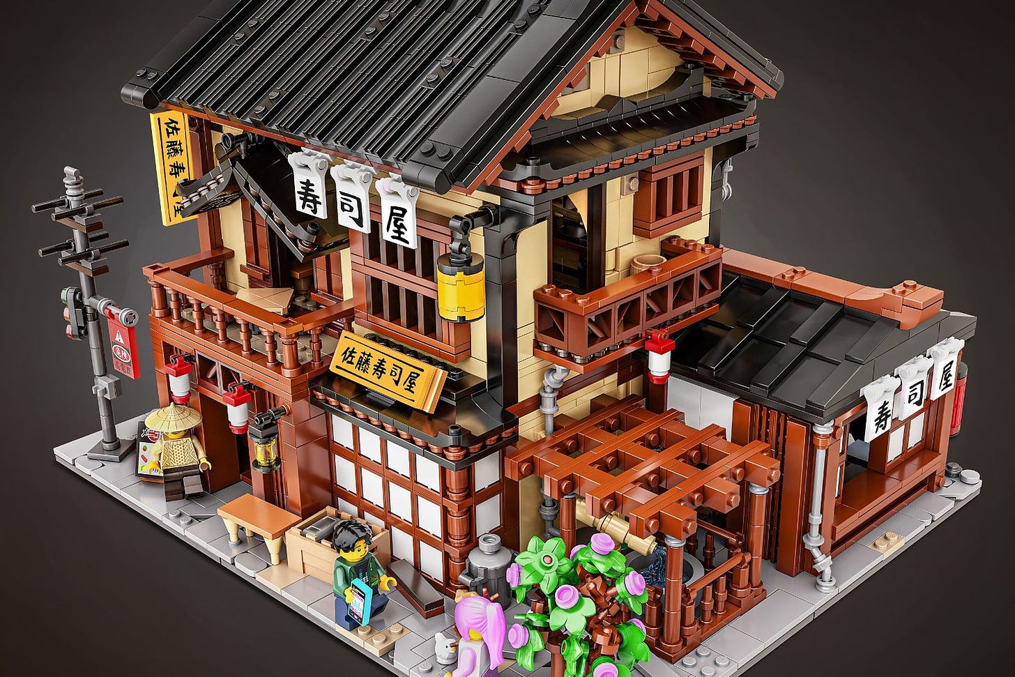 A traditional looking Japanese building made entirely out of Lego, with a fountain, flowers, two stories, entranceways and signs to the Sushi restaurant.